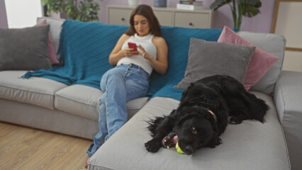 A young hispanic woman in casual attire sits on a living room sofa looking at her phone, while a...