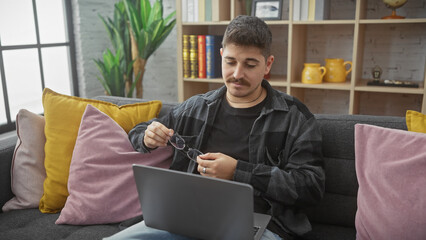 A casual hispanic man cleaning glasses while sitting on a couch in a cozy living room with a laptop.