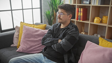 A thoughtful hispanic man with a moustache sits indoors with crossed arms on a sofa, surrounded by...