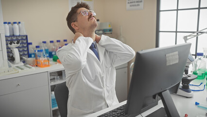 Young man in a lab coat rubbing neck pain at a workstation with microscope and computer in a...