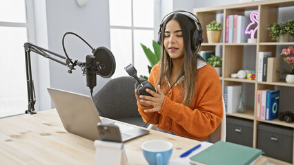 Hispanic woman podcaster in studio with microphone and laptop preparing to record content.