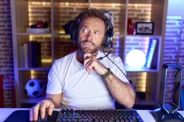 Middle age man with beard playing video games wearing headphones with hand on chin thinking about...