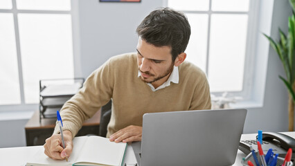 Handsome hispanic man working in a modern office environment, focused on writing in a notebook...