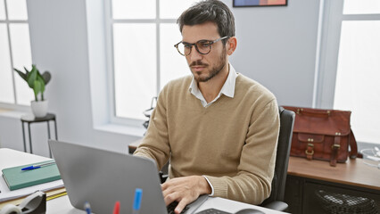 Hispanic man working on laptop in modern office, portraying professionalism with stylish eyewear...