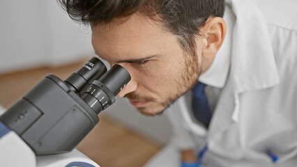 A young hispanic man in a lab coat intently using a microscope in an indoor laboratory setting.