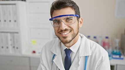 Handsome hispanic man with beard in lab coat and safety glasses inside laboratory smiling at camera.