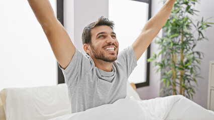 Smiling hispanic man stretching arms in bright bedroom interior