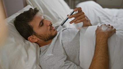 A young man lying in bed, checking a thermometer while appearing ill, provides a realistic...