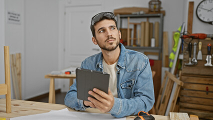 Handsome hispanic man in denim jacket pensively holding tablet in a carpentry workshop.