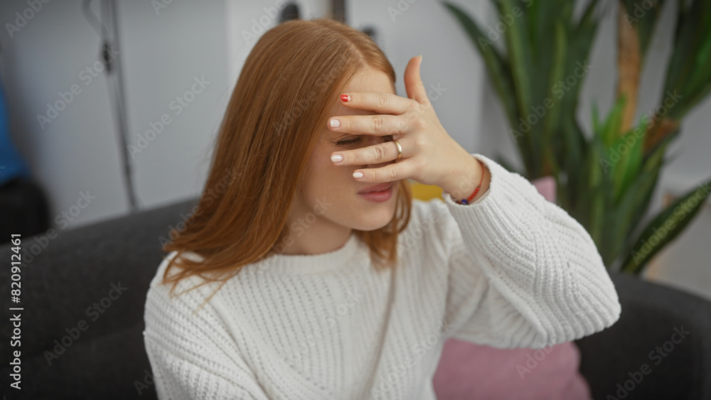 Sticker a young redhead woman in a white sweater covering her face with hand sitting in her living room