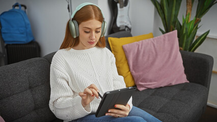 Obraz premium A young caucasian woman enjoys music with headphones in her apartment, using a tablet while seated on a sofa.