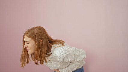 A young adult woman with red hair expressing stomach pain, isolated against a pink background