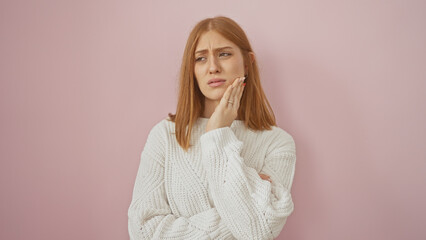 A young caucasian woman with red hair, in white sweater, touches her cheek in discomfort against a...