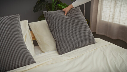 A hand adjusts a gray pillow on a neatly made bed in a sunlit bedroom with modern decor.