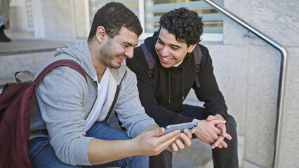 Two smiling men sitting on steps outdoors in a city, sharing a smartphone.