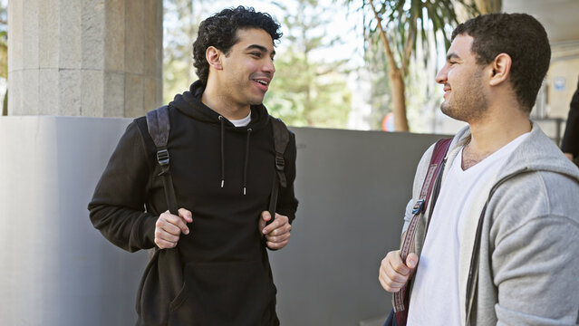 Two hispanic men chat amicably on a sunny urban street, portraying friendship and casual outdoor city life.