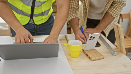 Two men collaboratively working in a carpentry workshop, using a laptop and taking measurements on...
