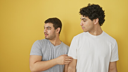 Two hispanic men in casual clothing stand against a plain yellow background, one touching the other's shoulder.