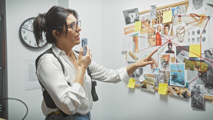 Middle-aged hispanic woman uses smartphone in detective office with evidence board