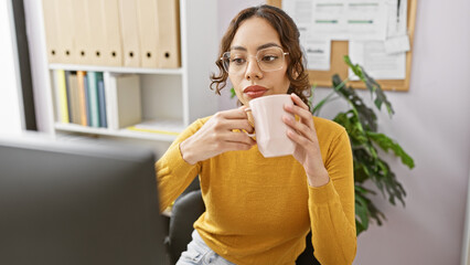 A professional woman in eyeglasses sips coffee at her office desk with a binder-filled shelf in the...