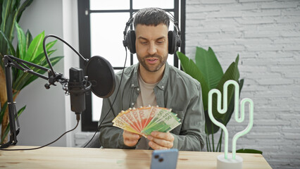 Hispanic man holding norwegian kroner in a radio studio, wearing headphones, with microphone and...