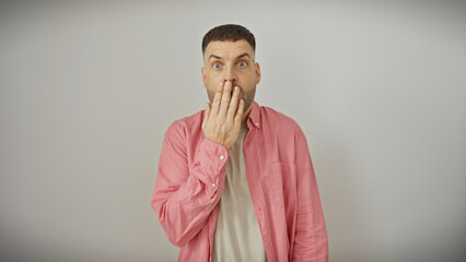 Surprised young man with a beard in a pink shirt covering his mouth against a white background