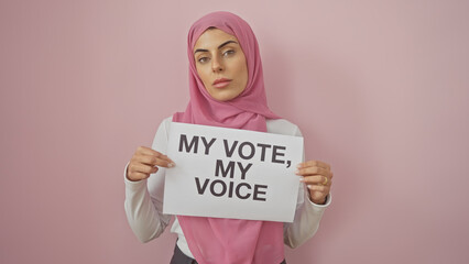 A confident young woman in a pink hijab holds a 'my vote, my voice' sign against an isolated pink...