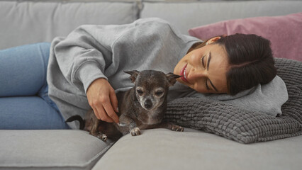Young hispanic woman affectionately petting a chihuahua on a couch in a cozy living room.