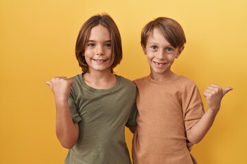 Two adorable boys, together, standing and hugging cheerfully; happily pointing their thumbs up with...