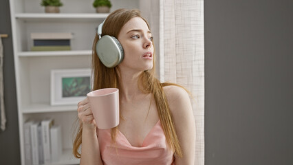 A pensive young woman wearing headphones holds a pink mug, gazing away in a modern bedroom.