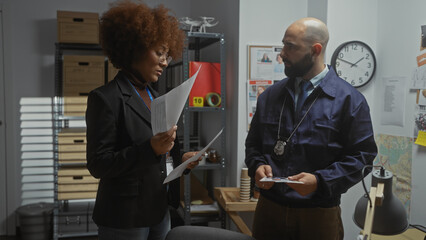 Man and woman detectives analyzing documents in a cluttered police station office.