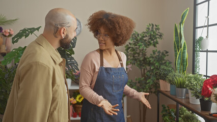 A woman florist in an apron conversing with a male customer amidst vibrant plants inside a cozy...