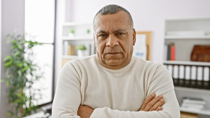 Middle-aged hispanic man with crossed arms looking serious in an office setting.