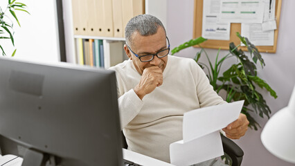 A thoughtful hispanic man reviews documents at his office desk, surrounded by indoor plants and...