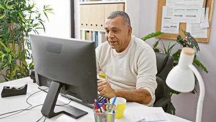 A middle-aged hispanic man focuses intently on his computer screen while seated at a cluttered desk...