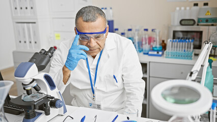 Hispanic man wearing lab coat and gloves analyzing samples with microscope in laboratory.
