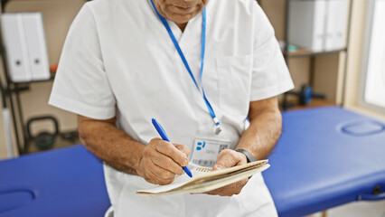 Middle-aged hispanic man in white lab coat writing notes in clinic room, showcasing healthcare professional at work.