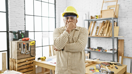 Pensive hispanic man in hardhat standing indoors at a carpentry workshop surrounded by tools and...