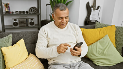 Obraz premium Smiling middle-aged hispanic man using a smartphone in a cozy modern living room