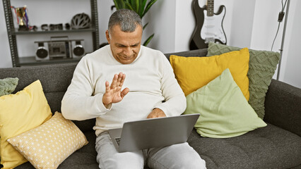 Smiling hispanic man using laptop in a cozy living room waves at screen, suggesting a video call at home.