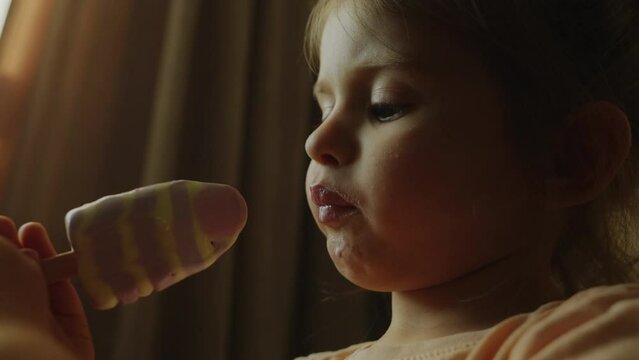 A little girl is happily eating an ice cream cone by a window