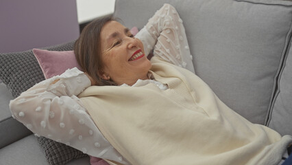 Smiling middle-aged woman relaxing on a gray sofa at home, conveying a sense of tranquility and...