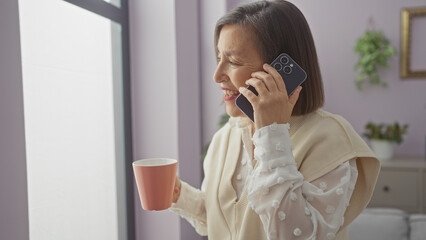 A smiling middle-aged woman enjoys a conversation on her smartphone while holding a pink mug in a cozy, pastel hued room at home.