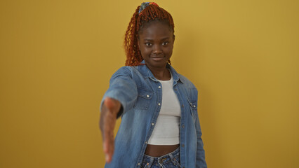 A confident young black woman in a denim outfit extends her hand against a vibrant yellow background.