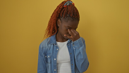 A young african american woman in a denim jacket rubbing her eyes against a yellow background.