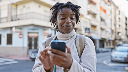 Smiling young woman with dreadlocks using smartphone on city street.