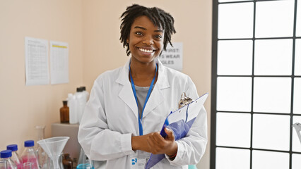 Smiling african american woman in a lab coat holding a clipboard in a laboratory setting.
