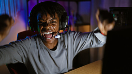 Excited african american woman wearing headphones celebrates a victory in a dark gaming room at...