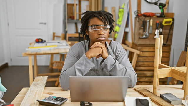 A thoughtful young woman with dreadlocks wearing safety glasses sits in a carpentry workshop with a laptop.