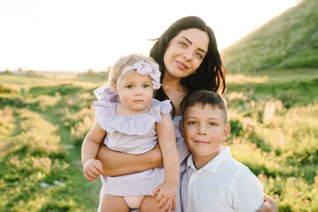 Mom hugs children standing in field at sunset. Portrait of daughter toddler and son hugging mother in nature on summer day. Concept of friendly family. Closeup. Mommy and kids spend time together.